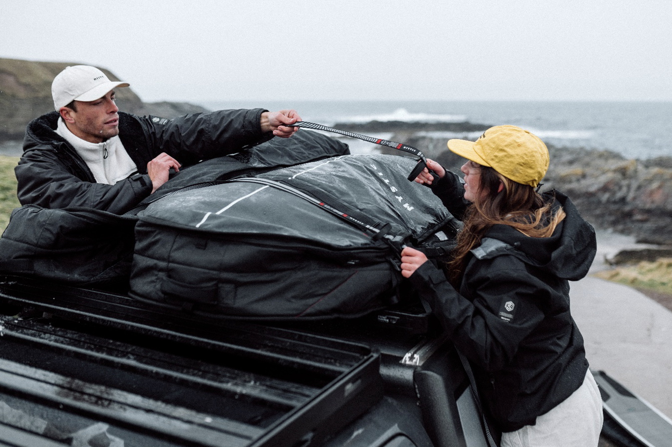 Zwei Surfer zurren eine große schwarze Boardbag auf dem Dachträger eines Autos am Meer fest