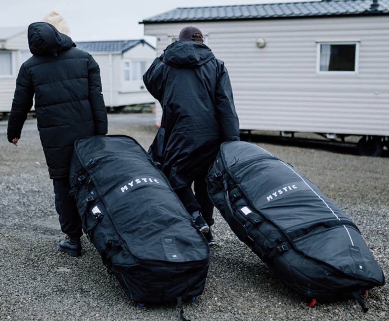 Zwei Reisende in Winterjacken ziehen große Mystic Boardbags über Kies an einem Mobilheim vorbei