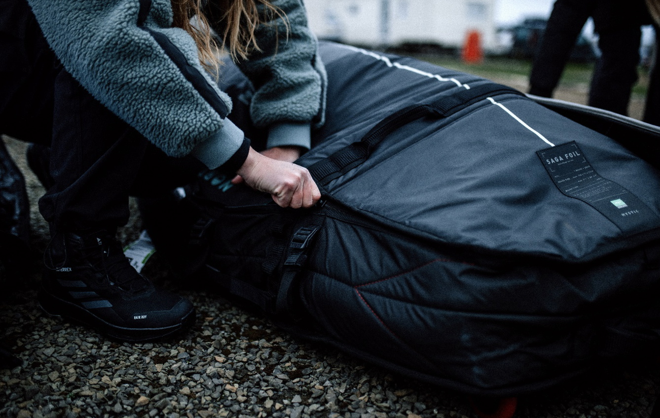 Reisender im Fleece kniet auf Kies und schließt den Reißverschluss einer großen dunkelblauen Boardbag