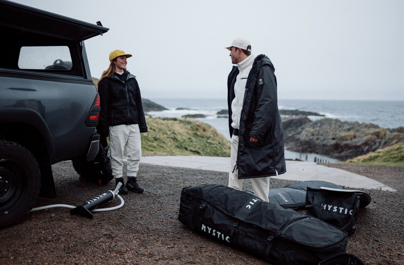 Zwei Surfer im Umziehmantel neben einem Geländewagen an der Küste, Mystic Boardbags am Boden