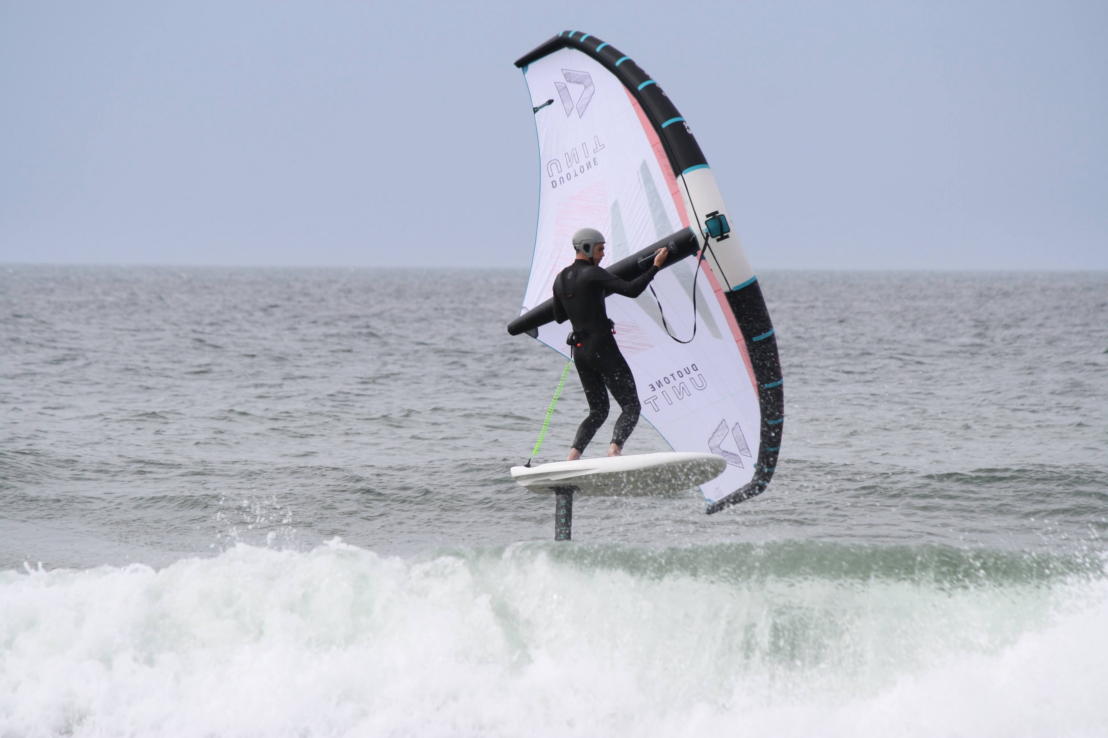 Wing foiler in a black wetsuit rising out of the whitewater on a foil board with a white wing