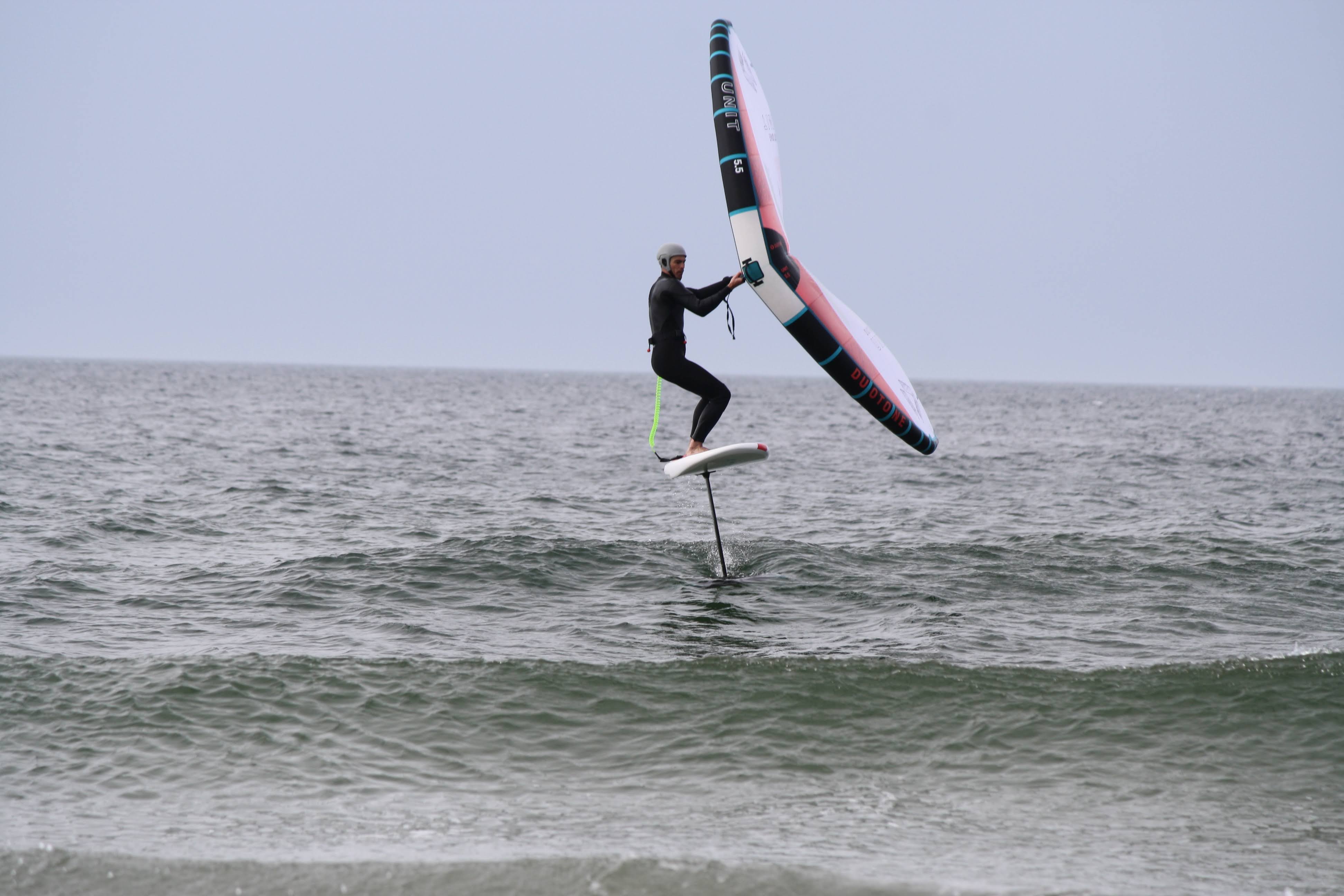 Wing foiler in a black wetsuit flying clear of the water holding a pink and black wing