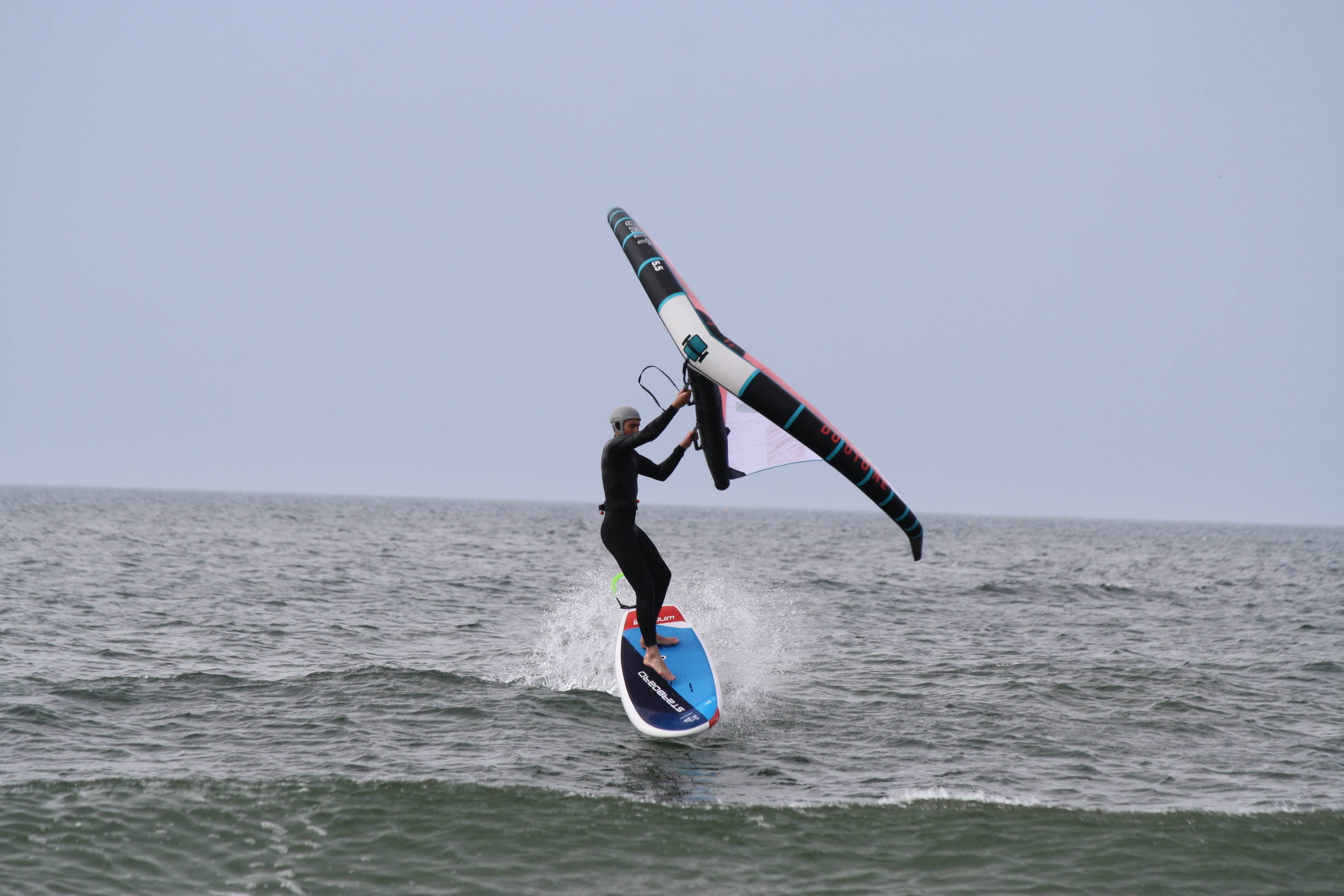 Wing foiler in a black wetsuit riding a blue and white board through the shorebreak