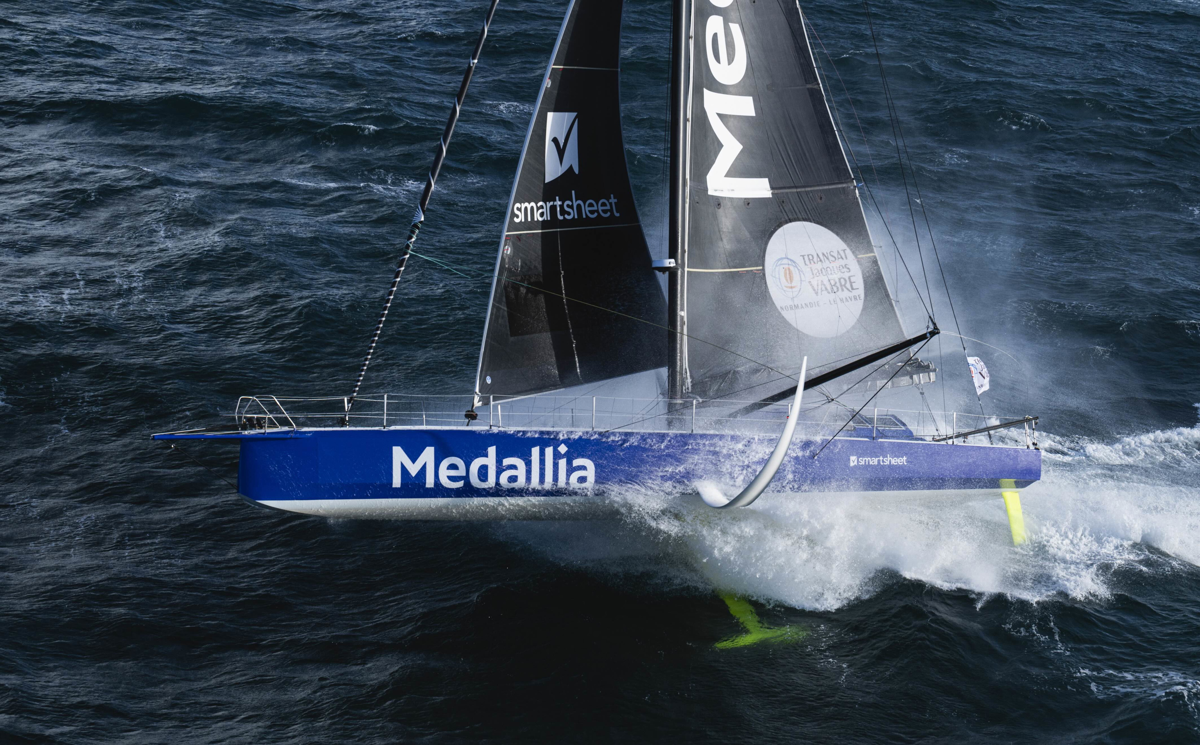 Blue and white IMOCA Medallia driving through waves on its foils with sponsor branding on the sail
