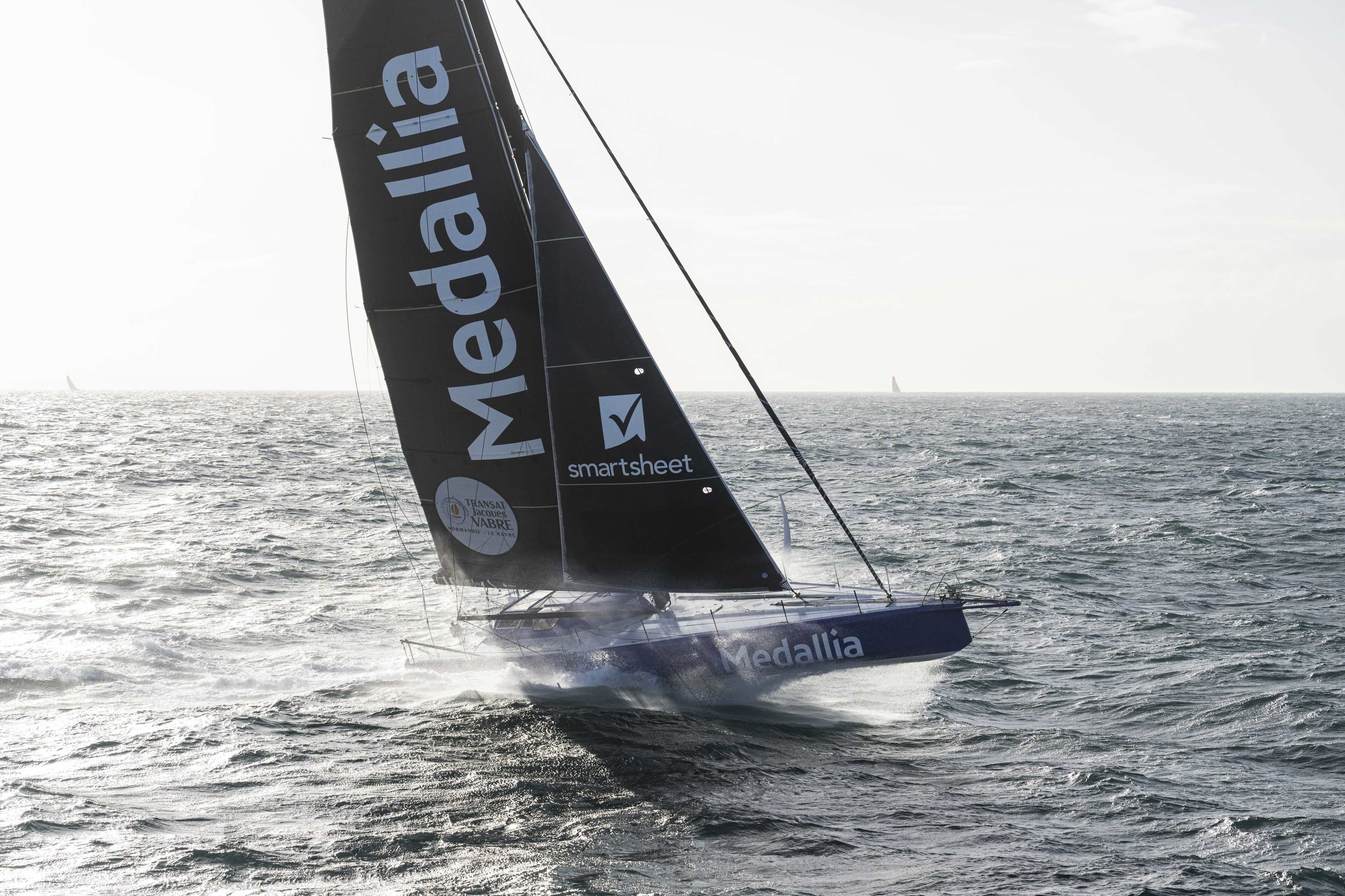 IMOCA racing yacht Medallia with black sails sailing under a grey sky on open ocean