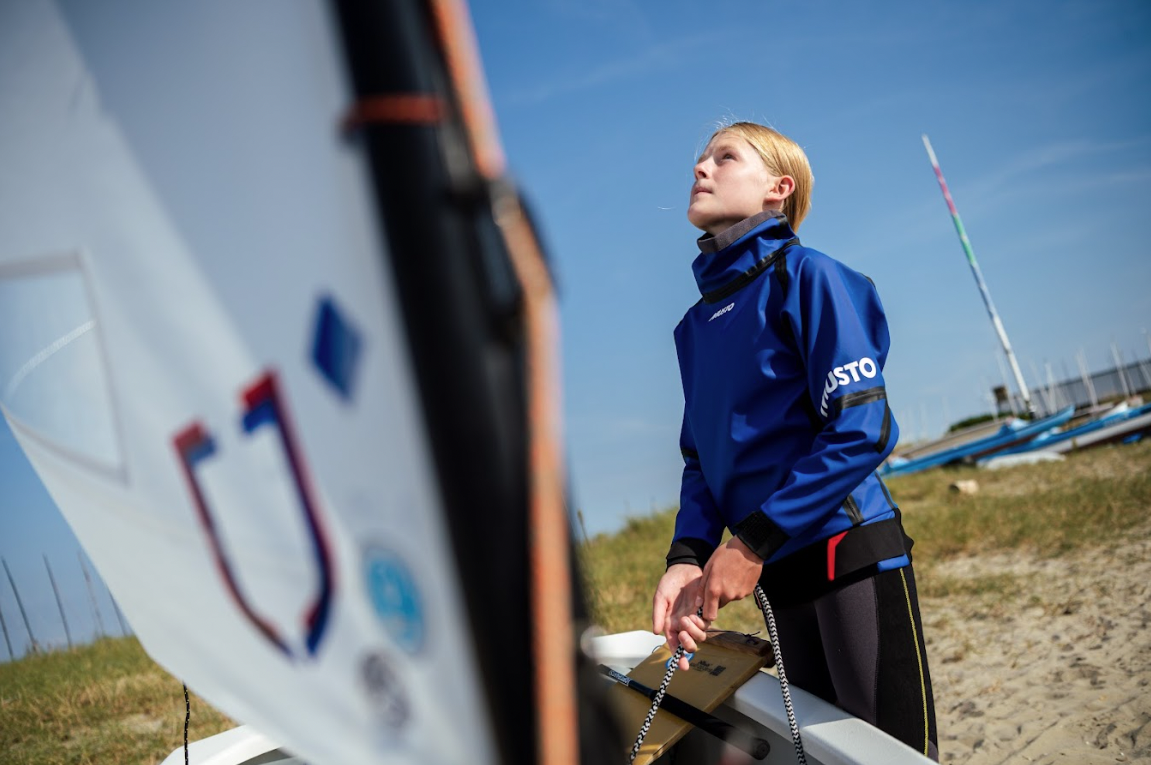 Young sailor in a blue sailing jacket rigging a dinghy on the shore of a sailing club