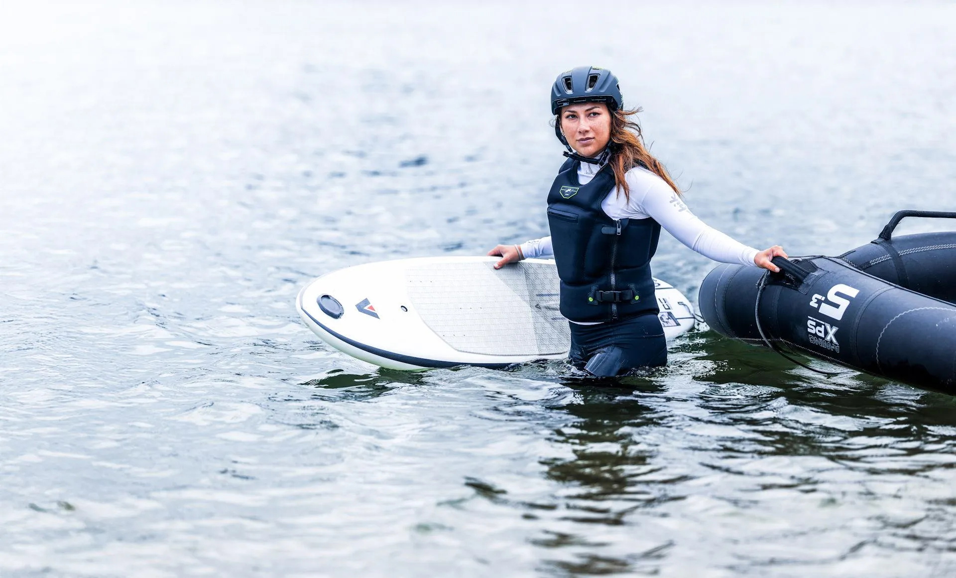 Sailor in a helmet and buoyancy aid standing waist-deep in the water beside a RIB, holding a board