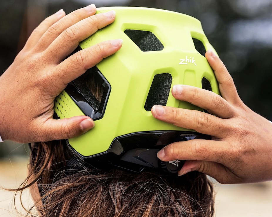 Hands fitting a fluorescent yellow vented Zhik watersports helmet onto someone's head