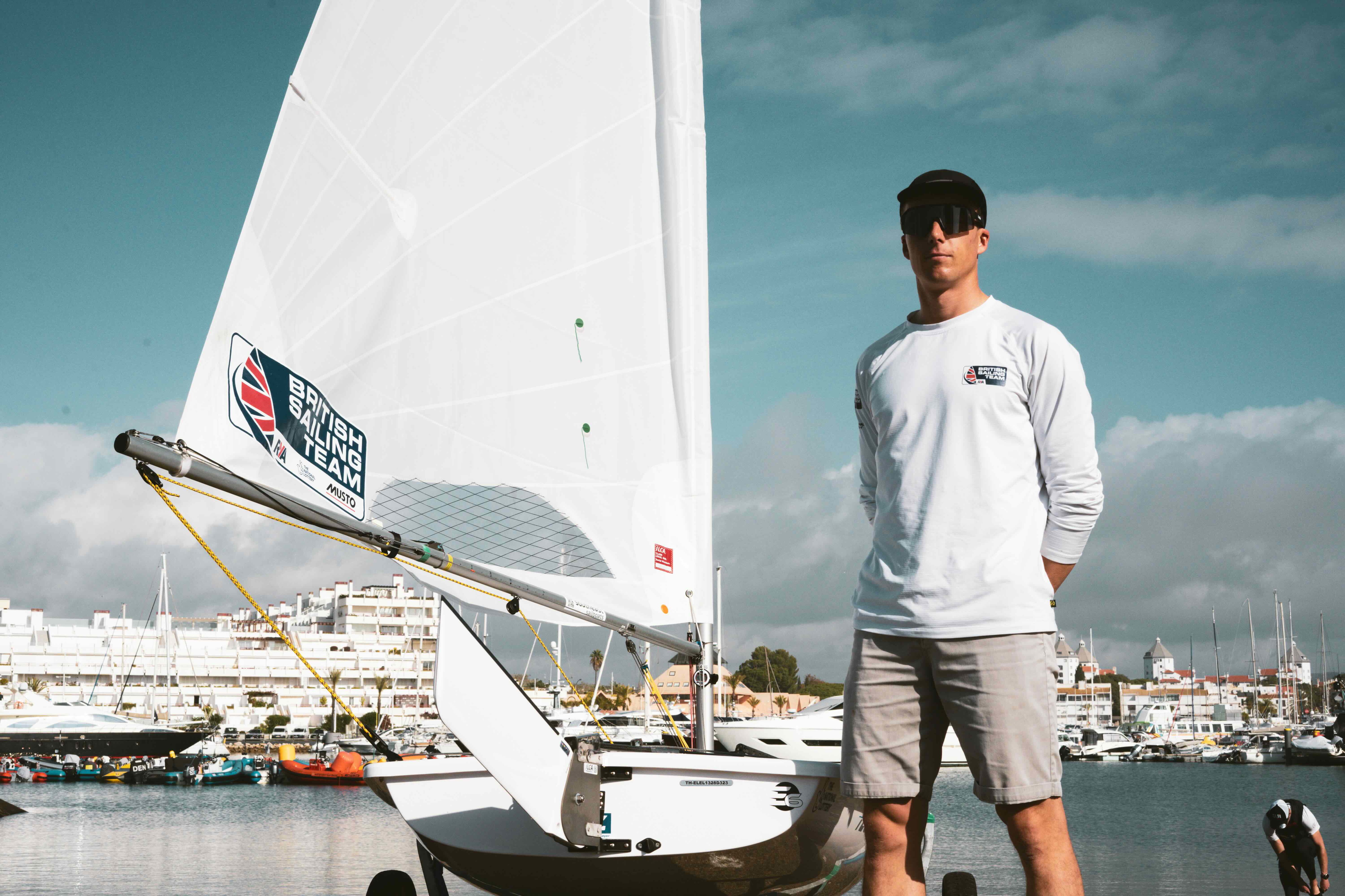 Young sailor in a white long-sleeved top and cap standing beside a British Sailing Team dinghy at a marina