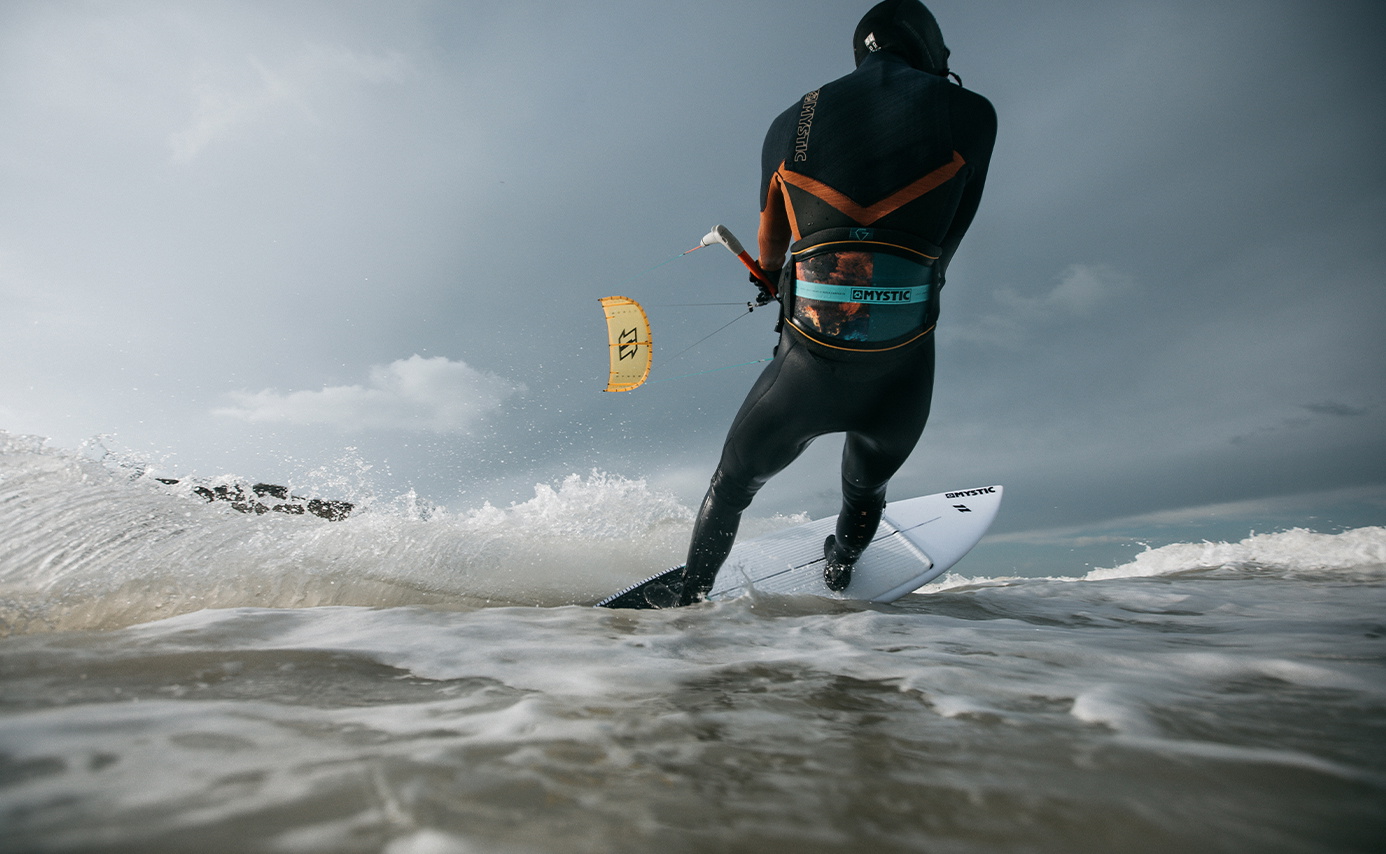 Kitesurfer im schwarzen Anzug mit Kapuze carvt auf dem Surfboard eine brechende Welle unter gelbem Kite