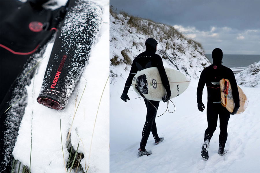 A rolled wetsuit lying in snow beside two surfers in black wetsuits carrying boards over snowy ground