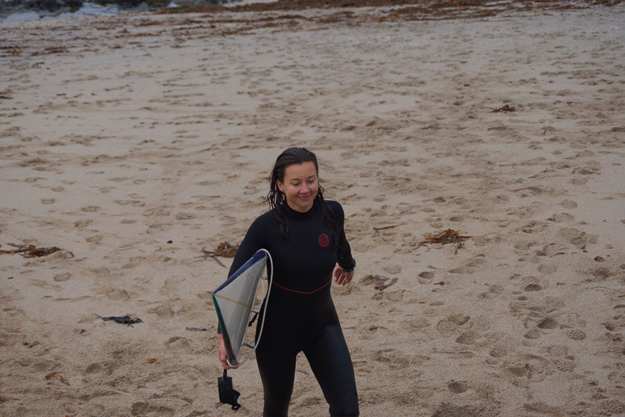 Smiling surfer in a black wetsuit walking up the sand with a board under her arm